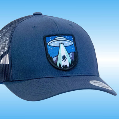 A navy blue trucker-style cap with a curved brim, mesh back panels, and a shield-shaped embroidered patch on the front featuring a UFO with a beam of light over mountains and a silhouetted figure of Bigfoot.