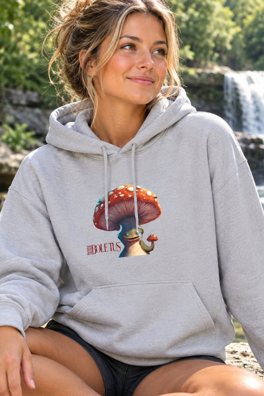 A woman wearing a light gray pullover hoodie with a colorful mushroom graphic and the word 'Boletus' printed on the chest, seated outdoors in front of a waterfall and greenery.