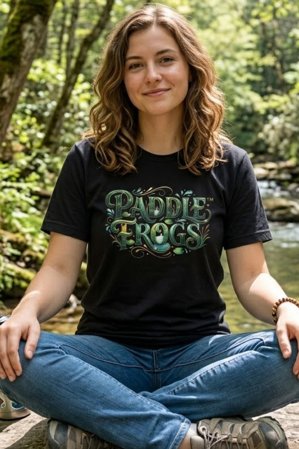 A woman sitting cross-legged outdoors by a stream wearing a black graphic paddleboarding t-shirt with 'PADDLE FROGS' in green and blue lettering, paired with blue jeans.