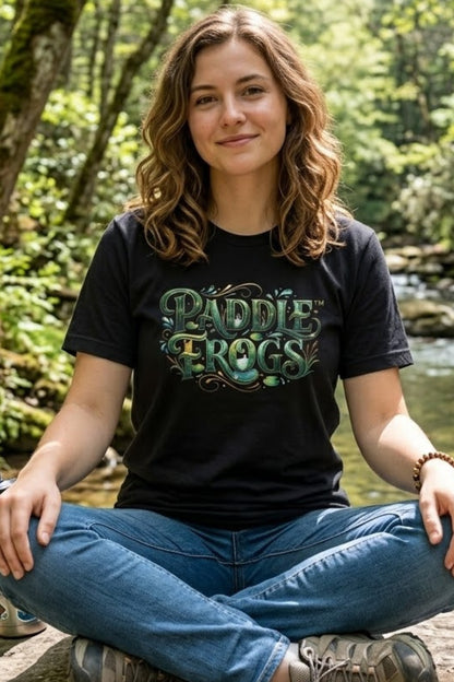A woman sitting cross-legged outdoors by a stream wearing a black graphic paddleboarding t-shirt with 'PADDLE FROGS' in green and blue lettering, paired with blue jeans.