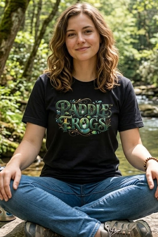 A woman sitting cross-legged outdoors by a stream wearing a black graphic paddleboarding t-shirt with 'PADDLE FROGS' in green and blue lettering, paired with blue jeans.