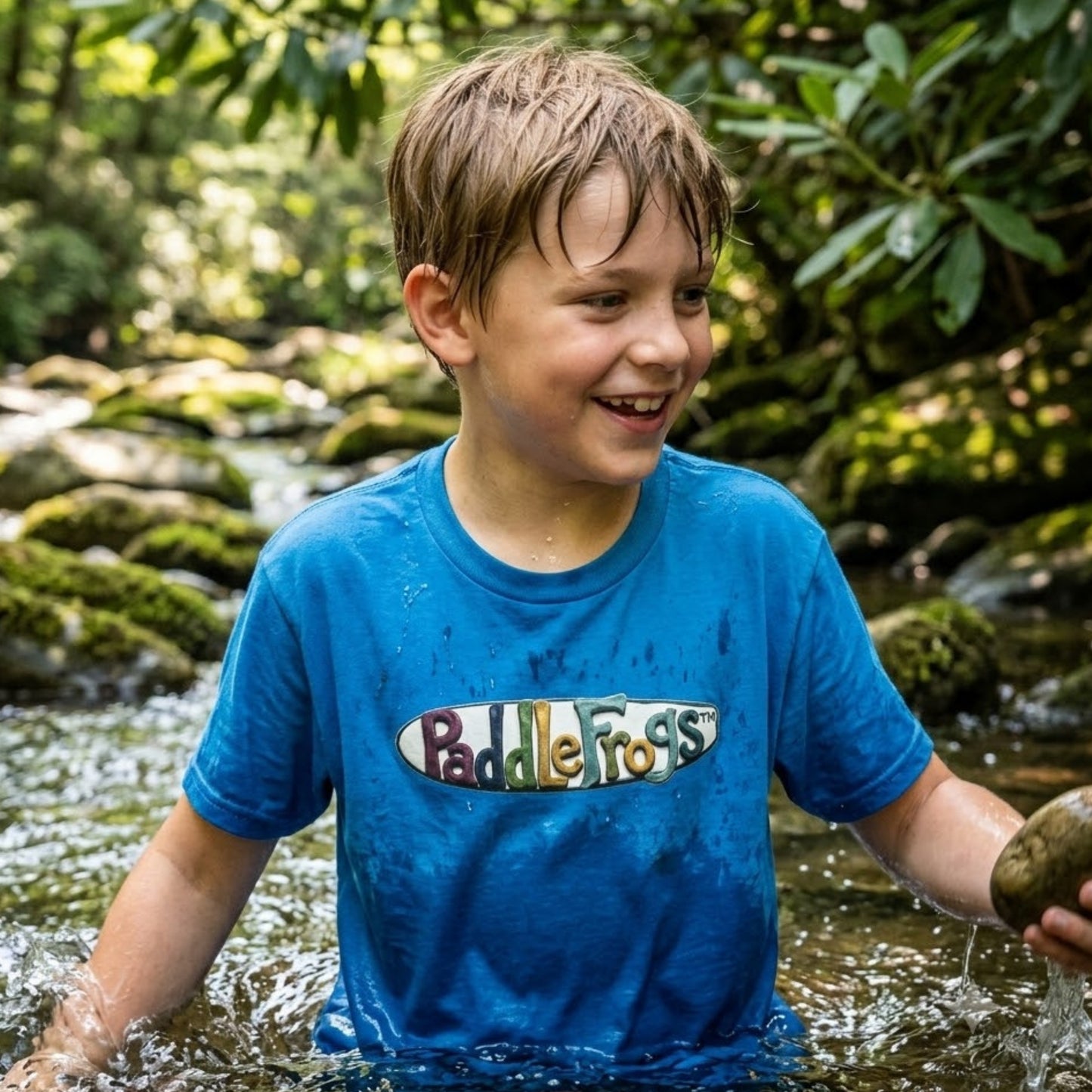 A boy wearing Paddle Frogs Kids bright blue Claymation SUP paddleboarding T-Shirt. He is in the creek playing with rocks.