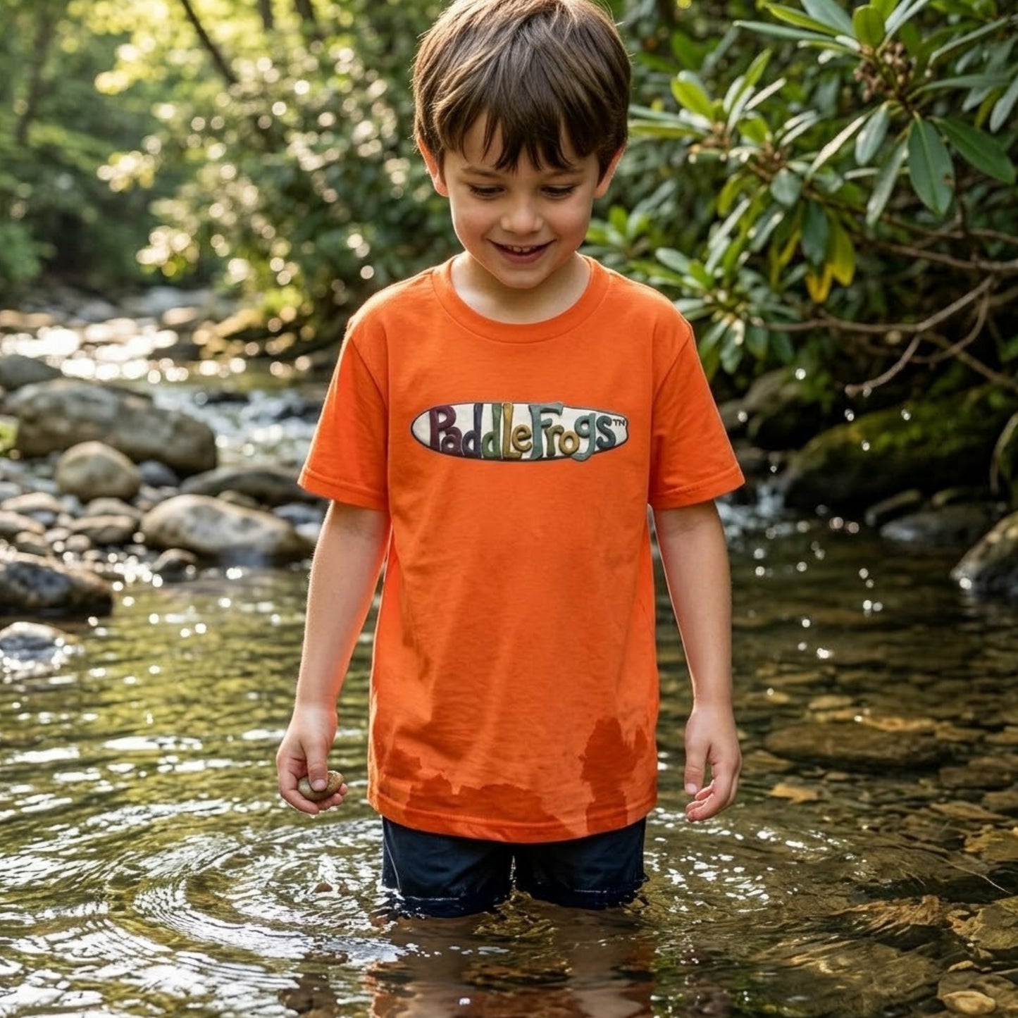 A young boy playing in a creek wearing an orange t-shirt by Paddle Frogs Kids. Claymation SUP paddleboarding tee.
