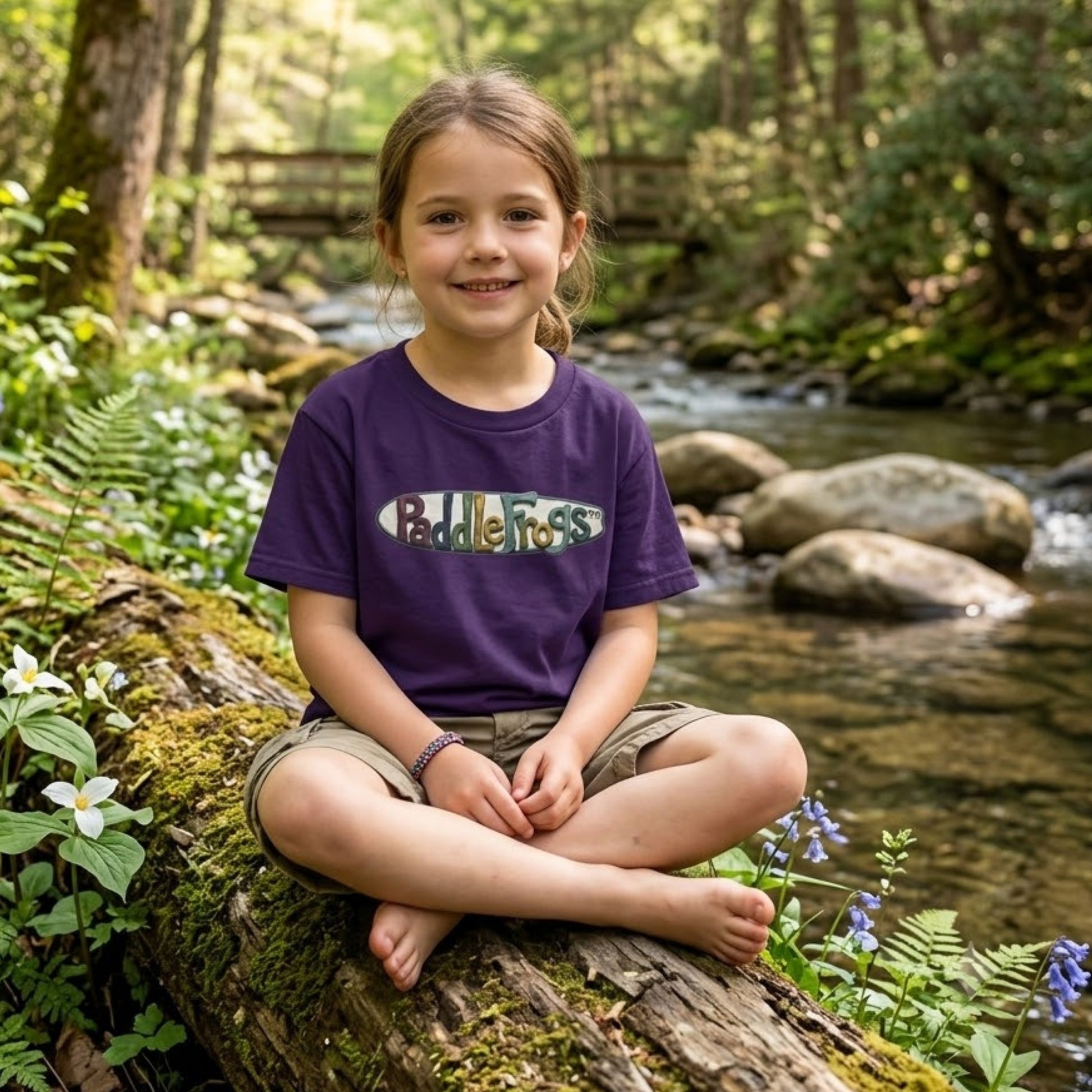 A young girl sits cross-legged on a mossy log beside a flowing forest stream, wearing a purple short-sleeve T-shirt Claymation paddleboard SUP Tee by Paddle Frogs kids