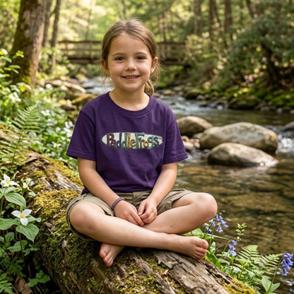 A young girl sits cross-legged on a mossy log beside a flowing forest stream, wearing a purple short-sleeve T-shirt Claymation paddleboard SUP Tee by Paddle Frogs kids