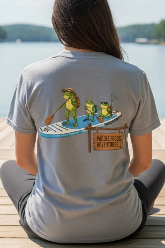 Rear view of a woman sitting on a wooden dock overlooking a lake, wearing a gravel grey t-shirt. The back of the shirt features a graphic of three frogs on a paddleboard next to a wooden sign that says 'Paddle Frogs Adventures'.