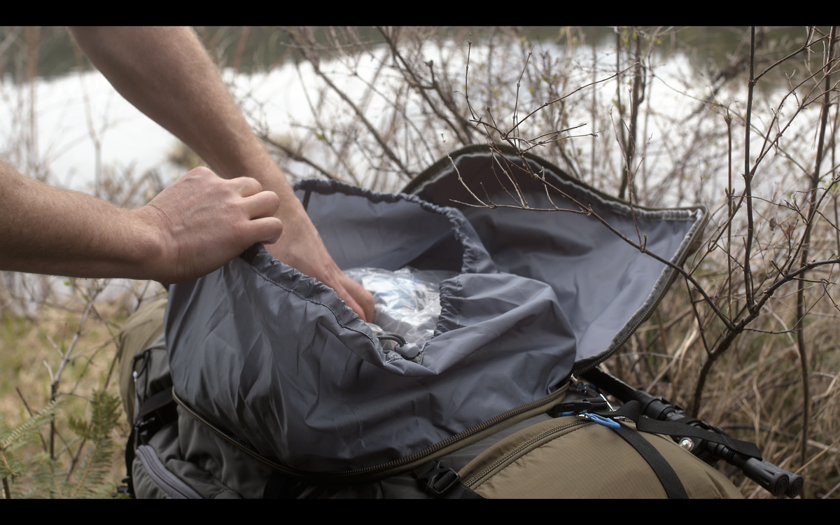 A close-up of a person's hands opening a gray waterproof pouch inside a tan and gray backpack, set outdoors near a body of water with dry brush in the background.
