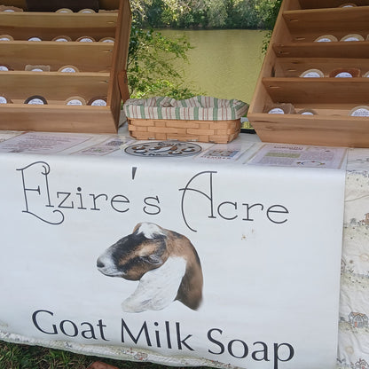 A handmade goat milk soap display table at an outdoor market, featuring a white banner with 'Elzire's Acre Goat Milk Soap' and a goat illustration, wooden trays of soaps on both sides, and a striped basket in the background. Located in Harriman, TN at the Paddle Frogs Sunday Market beside the Emory River & vendors have shade.