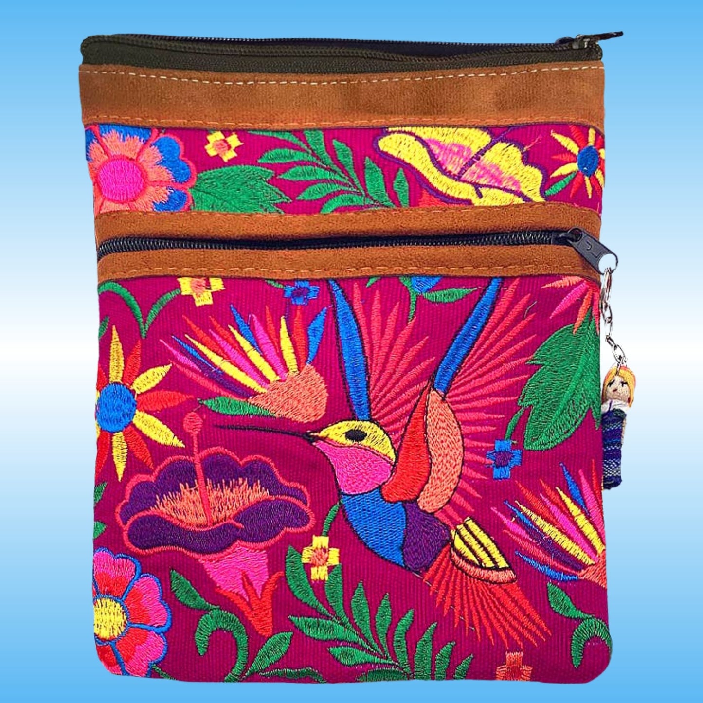 A small zippered two pouch crossbody purse with a vibrant pink and magenta embroidered design featuring a hummingbird, flowers, and tropical leaves, accented with brown faux-leather trim at the top and 2 black zippers. A worry doll included.