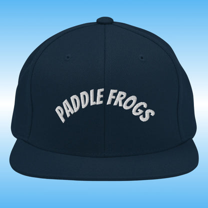 A dark navy blue flat-brim cap with 'PADDLE FROGS' embroidered in white arched text across the front, set against a light blue gradient background.