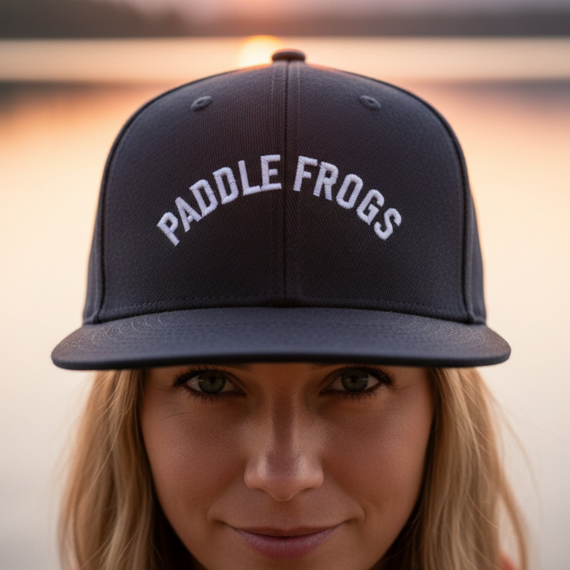 A close-up lifestyle shot of a woman wearing a dark blue snapback cap with 'PADDLE FROGS' embroidered in white arched text on the front, set against a warm, blurred sunset background.