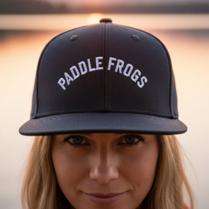 A close-up lifestyle shot of a woman wearing a dark blue snapback cap with 'PADDLE FROGS' embroidered in white arched text on the front, set against a warm, blurred sunset background.