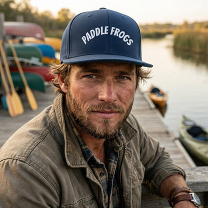 A man wearing a navy blue Paddle Frogs baseball cap and an olive green jacket, leaning on a wooden dock next to a calm lake with canoes in the background.