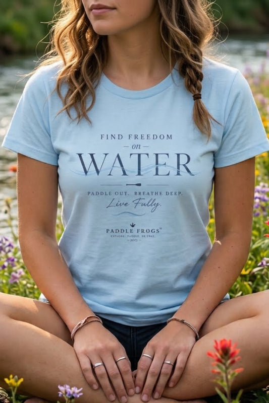 A woman sitting outdoors by a body of water wears a light blue graphic T-shirt with the words 'FIND FREEDOM ON WATER' and smaller text 'Paddle Out, Breathe Deep, Live Fully' plus the Paddle Frogs logo.
