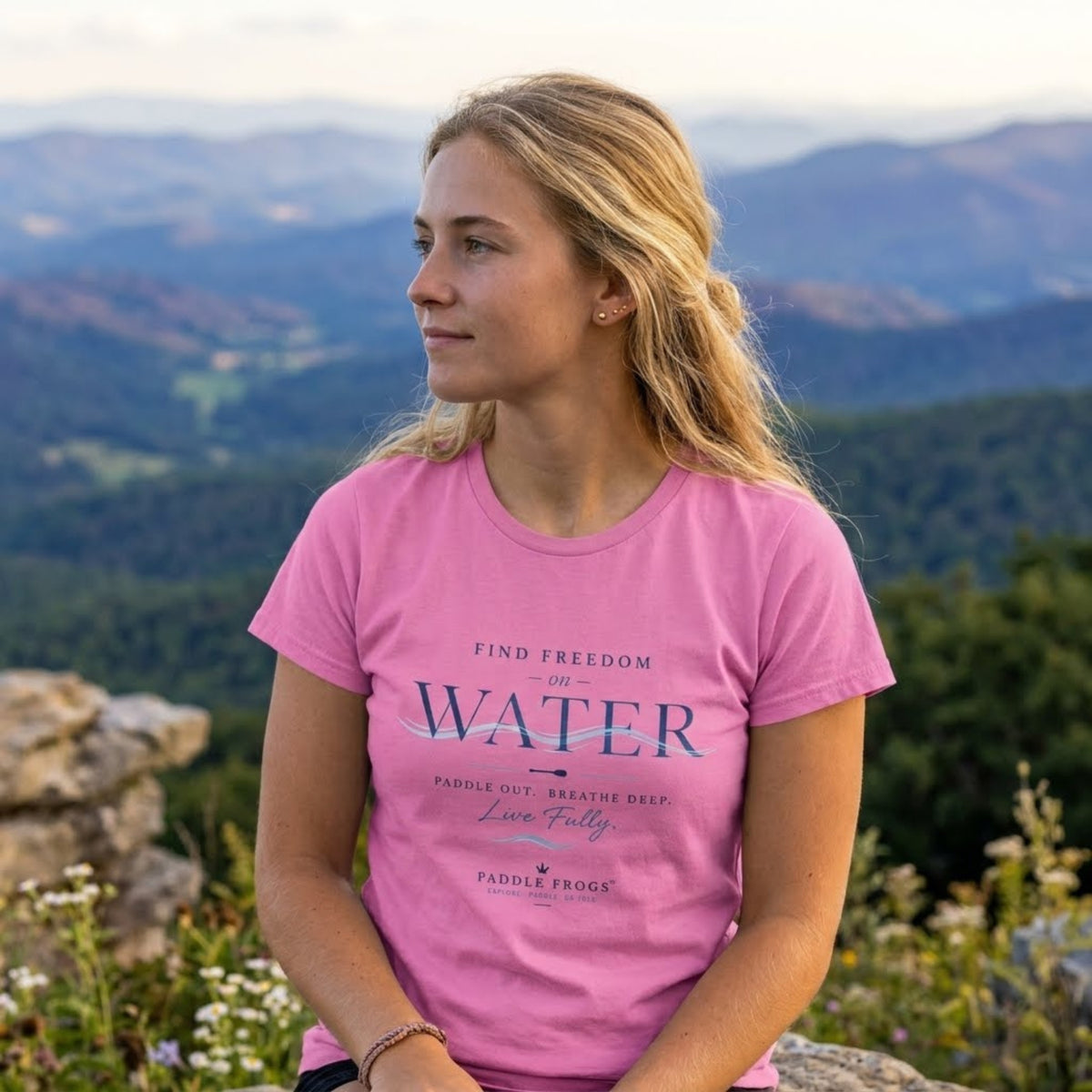 A woman wearing a pink graphic paddleboarding t-shirt with the text 'FIND FREEDOM ON WATER' is pictured outdoors with a scenic mountain landscape in the background.