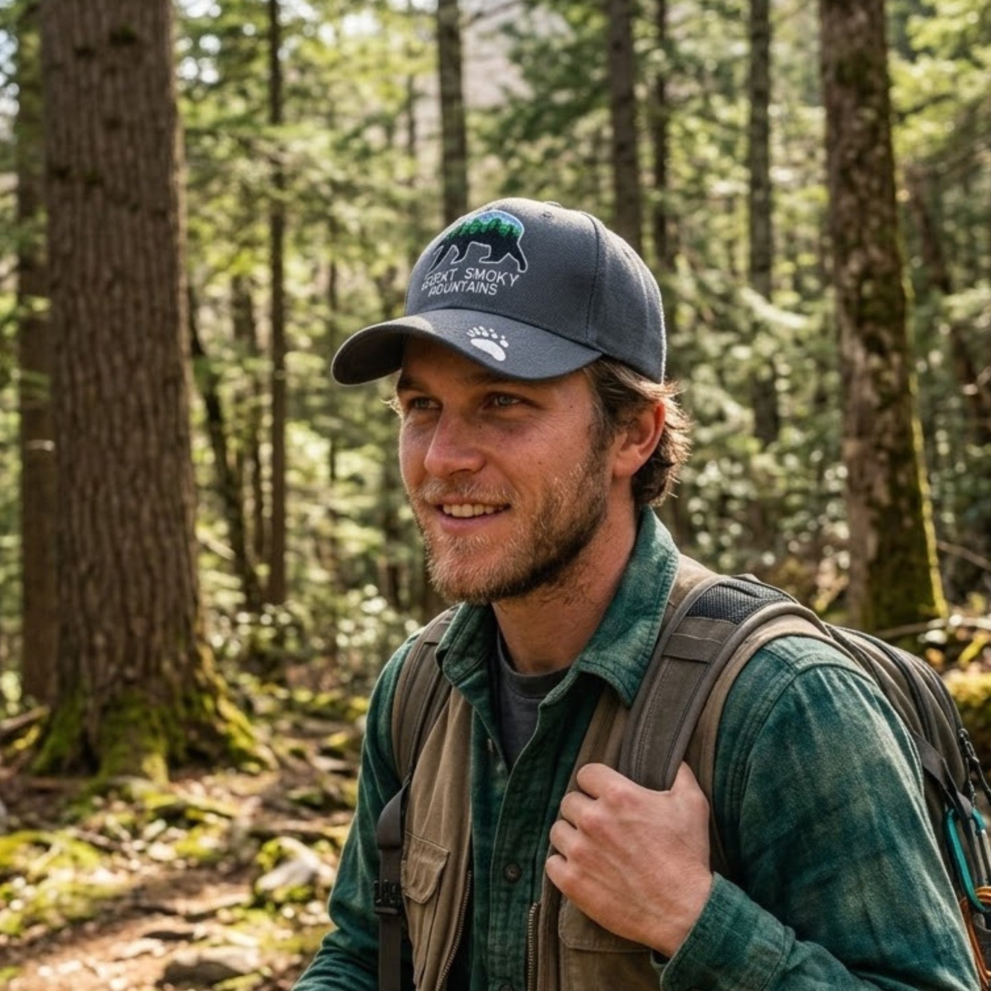 A man wearing a dark gray baseball cap with a green and white 'black bear Wilderness' logo, a green flannel shirt, and a backpack, standing in a forest.