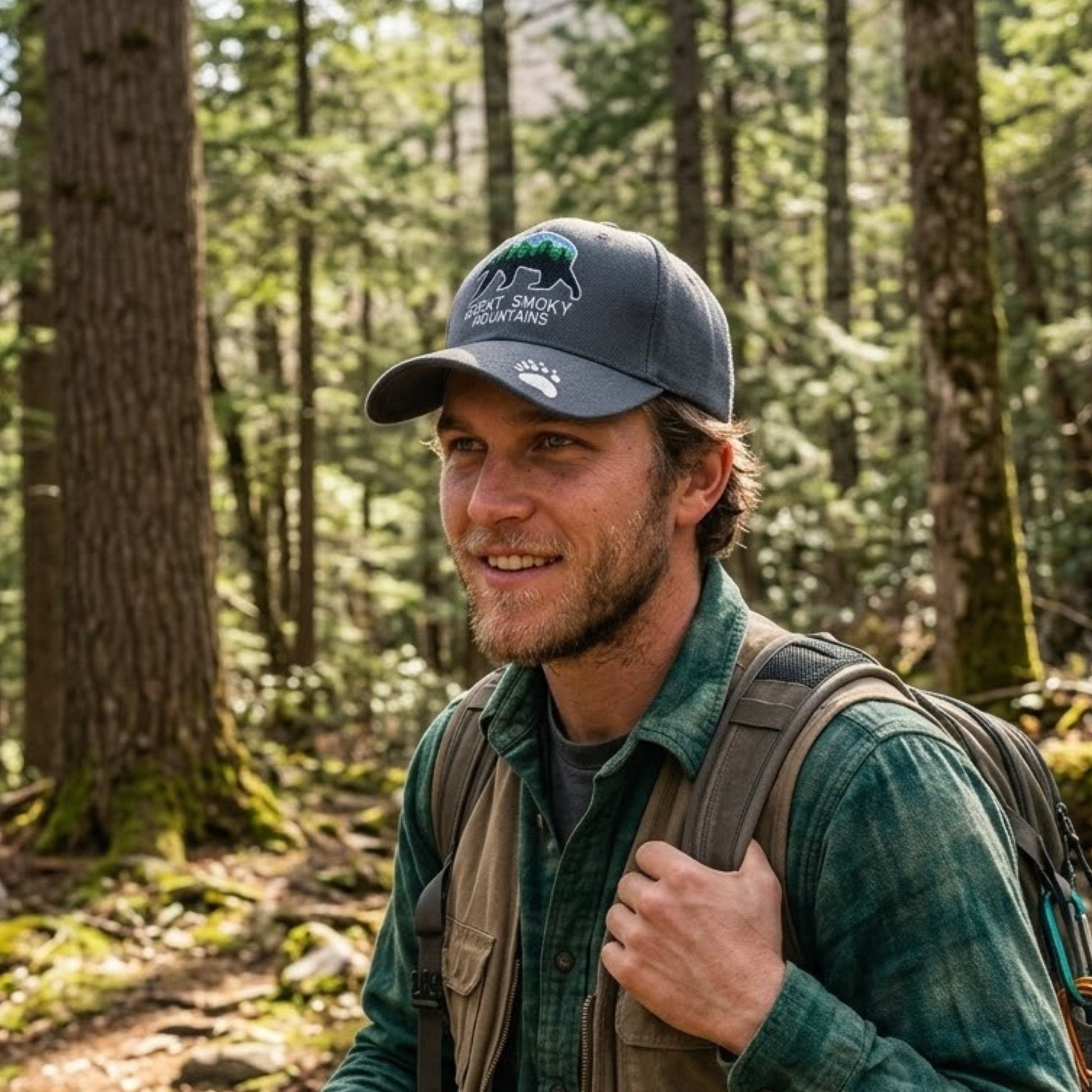 A man wearing a dark gray baseball cap with a green and white 'black bear Wilderness' logo, a green flannel shirt, and a backpack, standing in a forest.