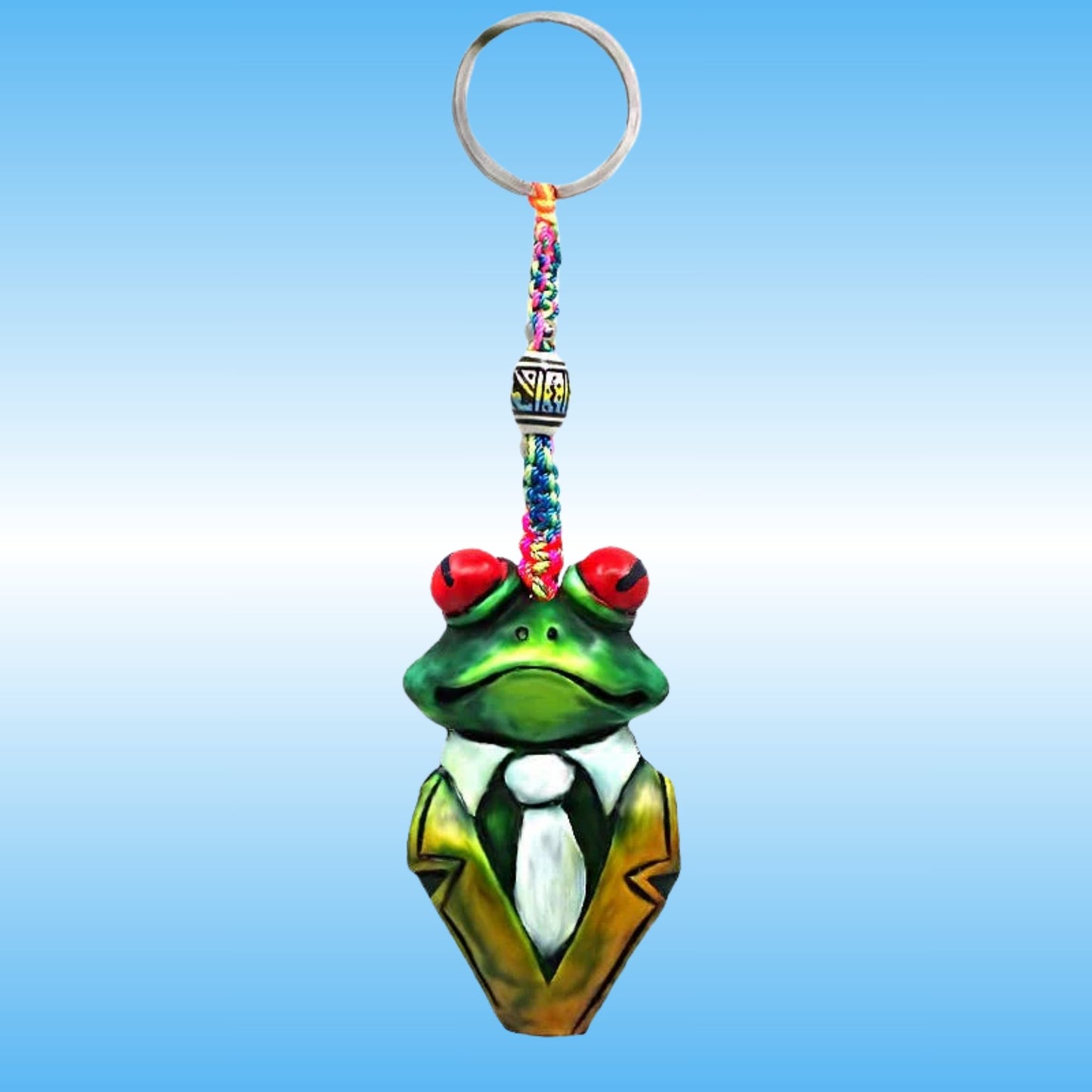 A frog-shaped keychain charm with a green body, red eyes, a white shirt, and a gold jacket, attached to a colorful braided strap and a silver key ring, shown against a blue gradient background.