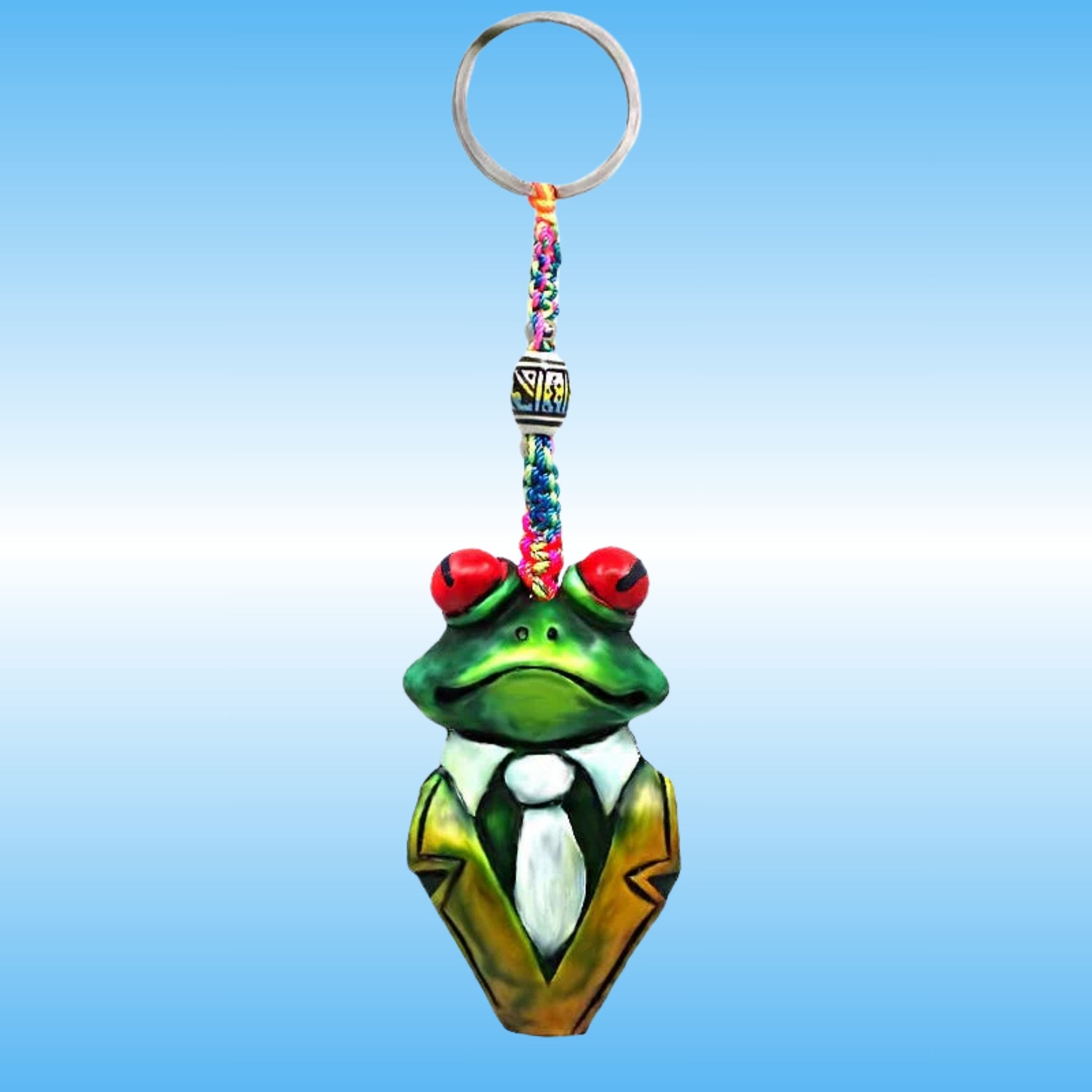 A frog-shaped keychain charm with a green body, red eyes, a white shirt, and a gold jacket, attached to a colorful braided strap and a silver key ring, shown against a blue gradient background.