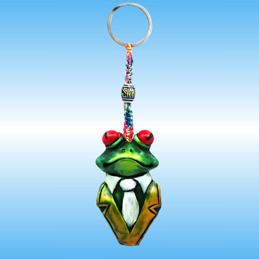 A frog-shaped keychain charm with a green body, red eyes, a white shirt, and a gold jacket, attached to a colorful braided strap and a silver key ring, shown against a blue gradient background.