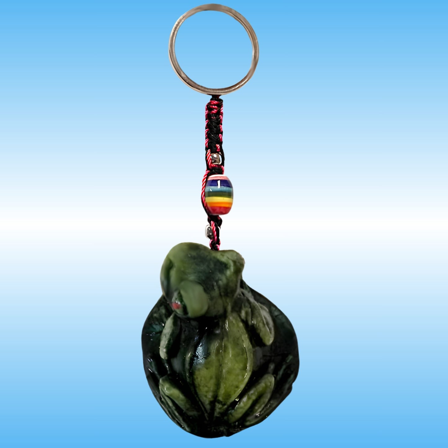 A handmade keychain featuring a carved green stone frog figurine, a red and black braided cord, a small rainbow-striped bead, and a silver metal key ring, set against a blue gradient background.