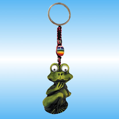 A green frog-shaped keychain charm with large cartoon Google eyes, sitting crouched and holding her hand near her face, attached to a metal key ring with a braided cord and a small rainbow-striped bead, shown against a blue gradient background.