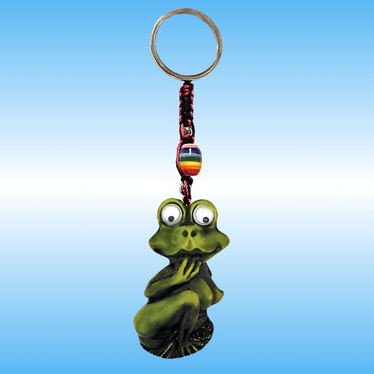 A green frog-shaped keychain charm with large cartoon Google eyes, sitting crouched and holding her hand near her face, attached to a metal key ring with a braided cord and a small rainbow-striped bead, shown against a blue gradient background.