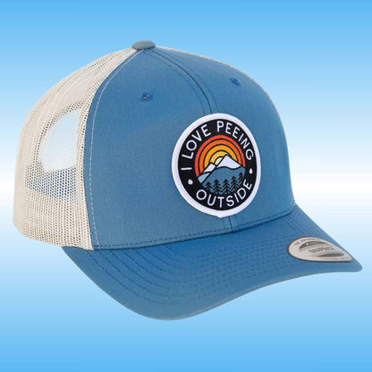 A blue and white trucker cap with a circular patch on the front that reads 'I LOVE Peeing OUTSIDE' with a mountain and rainbow graphic. The cap has a blue front panel and brim, white mesh back, and a silver visor sticker.