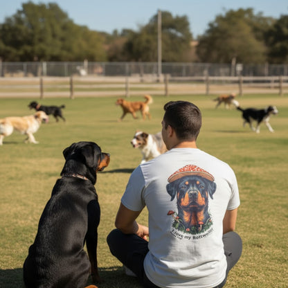 Man sitting on grass with a dog, wearing a t-shirt with a Rottweiler design, at a park with other dogs in the background.