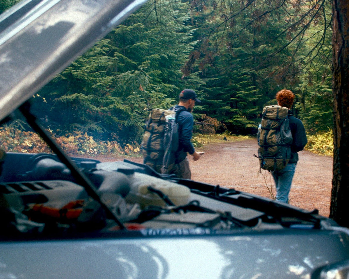 Two people with large hiking backpacks walk away from a vehicle on a forest road, with trees and brush in the background.