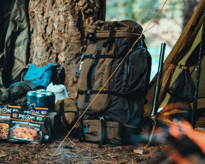 A large brown and black technical backpack stands next to a tent at a forest campsite, with PEOK snack boxes, water bottles, and camping gear visible in the scene.