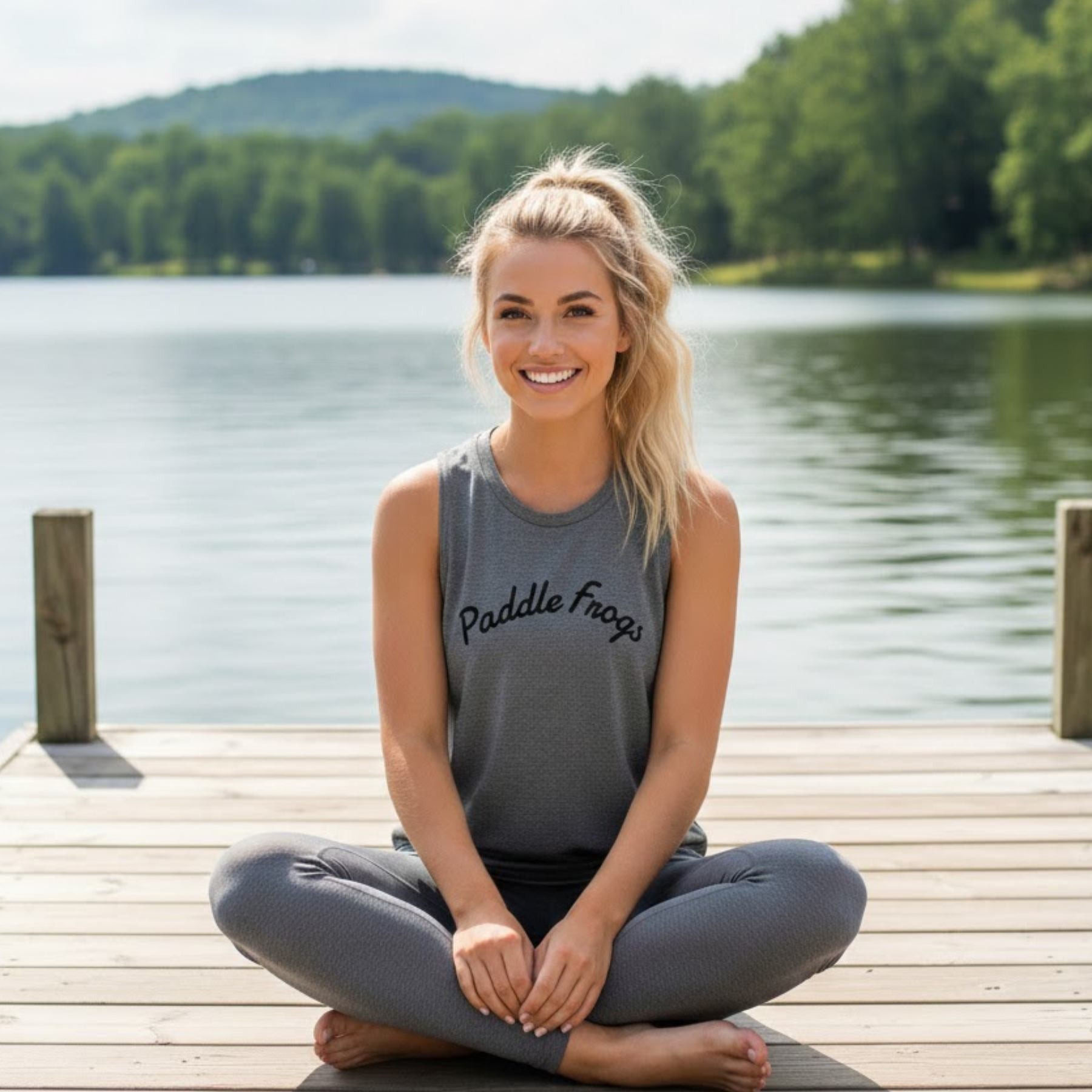 A healthy blonde model sitting crisscross on a lakeside pier wearing the grey Serene Stream Tank, showcasing the soft drape and comfortable lifestyle fit of the Paddle Frogs muscle tee.