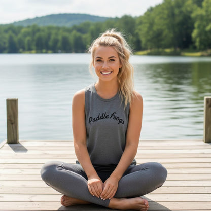 A healthy blonde model sitting crisscross on a lakeside pier wearing the grey Serene Stream Tank, showcasing the soft drape and comfortable lifestyle fit of the Paddle Frogs muscle tee.