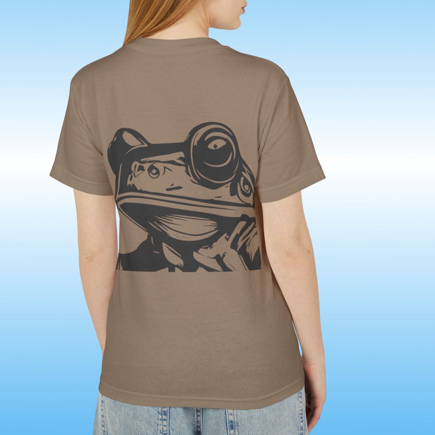 Lady  wearing a brown t-shirt with a frog graphic on the back against a blue gradient background Paddle Frogs Earthy T-Shirt Collection featuring the Frog face on the backside