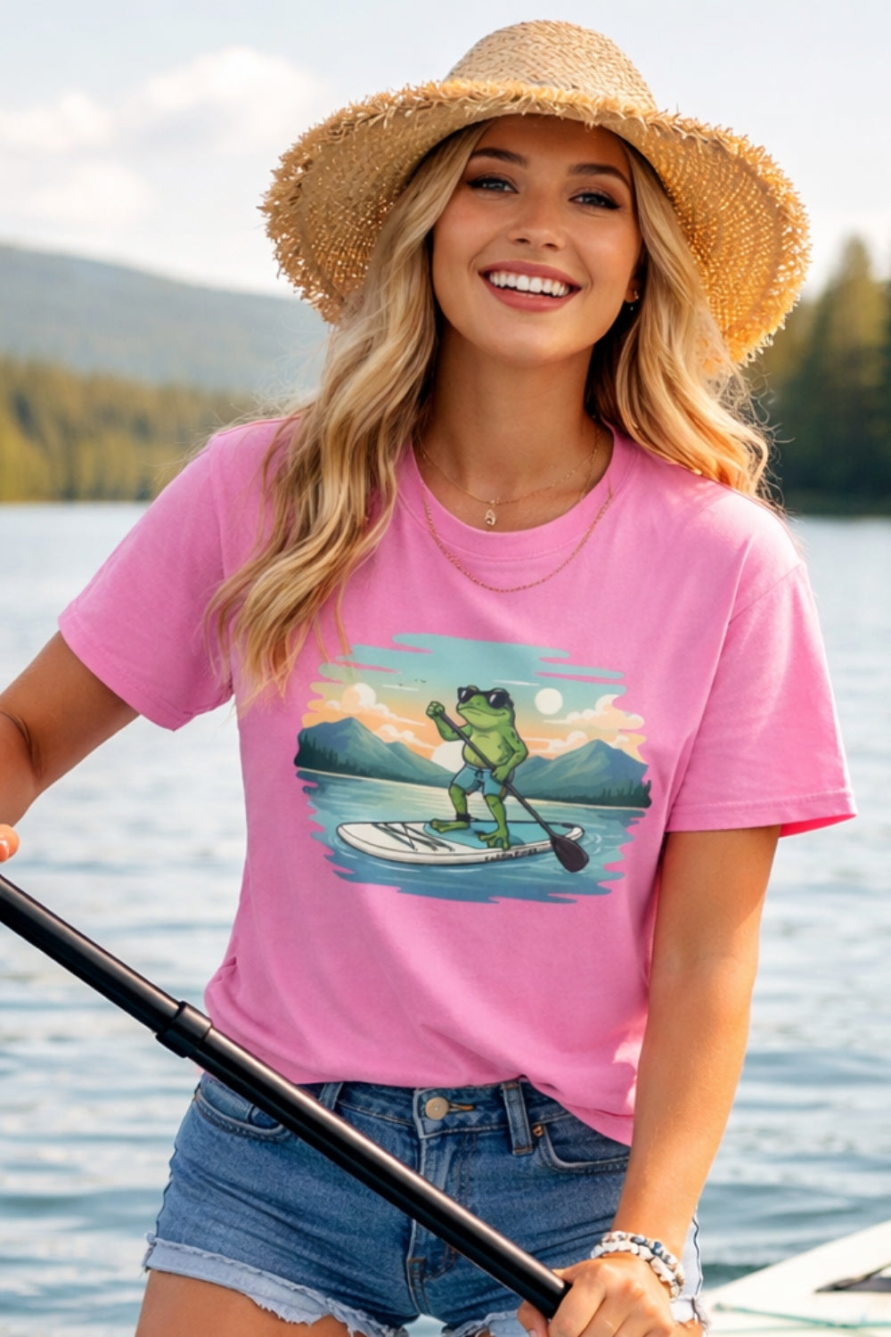 A blonde woman in a straw hat and denim shorts smiling on a lake while holding a paddle. She is wearing an azalea pink t-shirt featuring a graphic illustration of a cartoon frog wearing sunglasses and paddleboarding on a calm lake with mountains in the background.