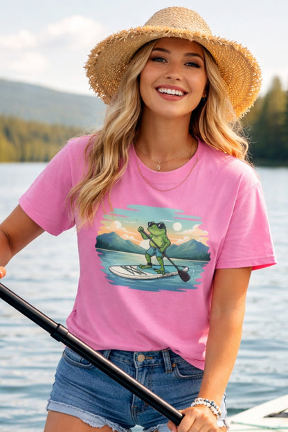 A blonde woman in a straw hat and denim shorts smiling on a lake while holding a paddle. She is wearing an azalea pink t-shirt featuring a graphic illustration of a cartoon frog wearing sunglasses and paddleboarding on a calm lake with mountains in the background.