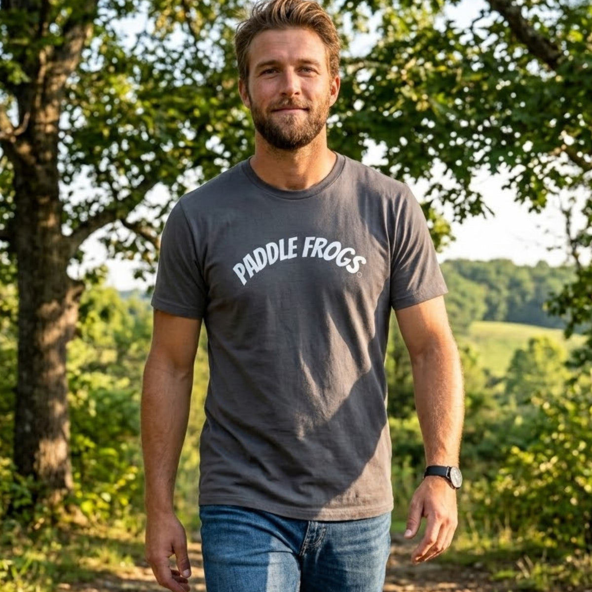 A man wearing a dark gray crewneck t-shirt with 'PADDLE FROGS' printed in white arched text across the chest, standing outdoors in a wooded setting.