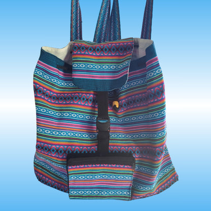 A colorful woven Peruvian backpack with a geometric multicolor pattern, featuring a front pocket and a black buckle strap closure, set against a light blue gradient background.