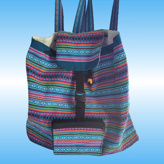 A colorful woven Peruvian backpack with a geometric multicolor pattern, featuring a front pocket and a black buckle strap closure, set against a light blue gradient background.