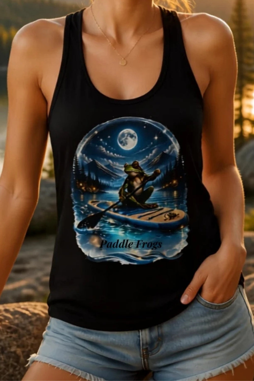 A woman wearing a black tank top with a detailed fantasy graphic of a frog paddleboarding under a full moon, posed outdoors in front of a mountain lake at sunset.