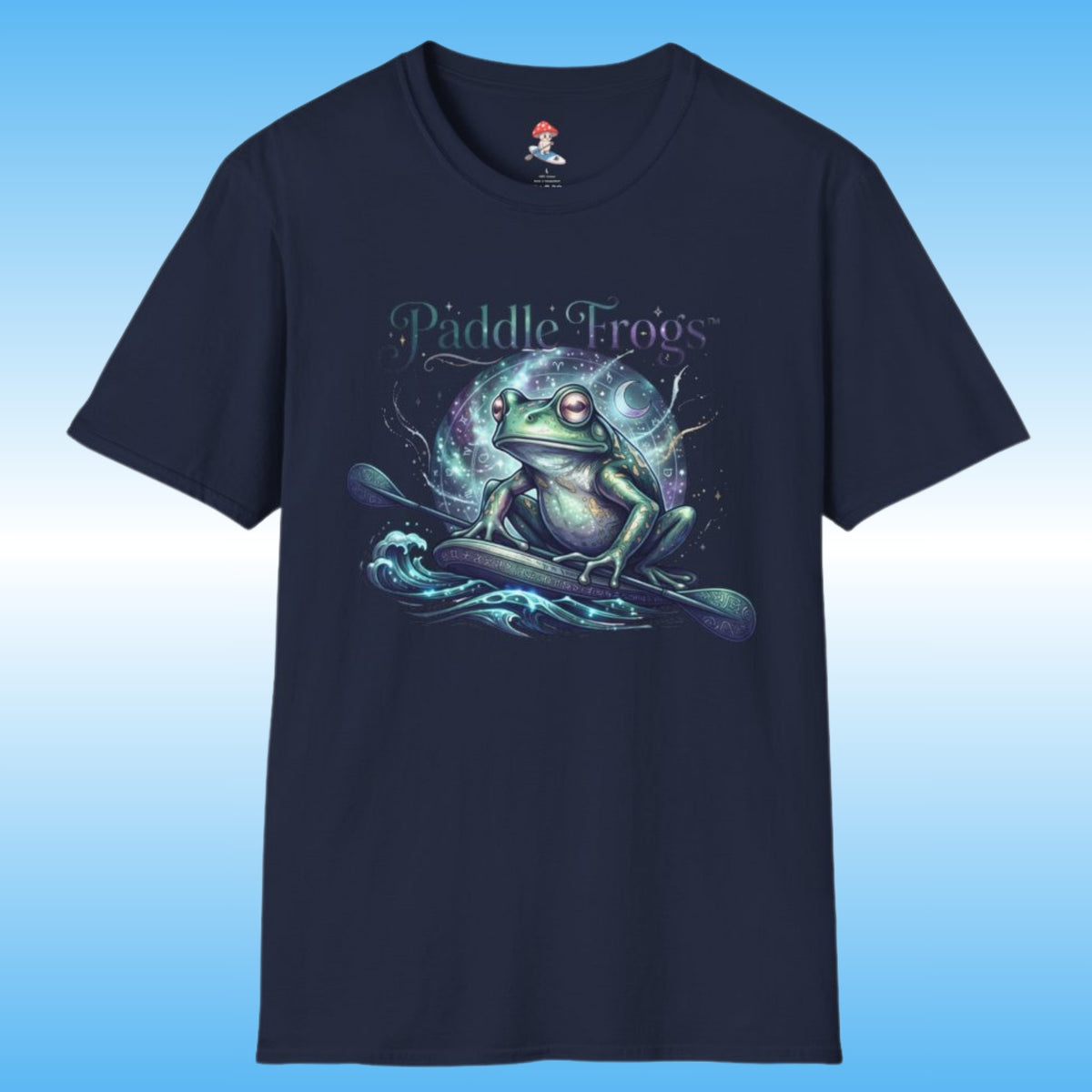 A navy blue short-sleeve t-shirt with a graphic of a green frog paddleboarding on stylized water waves, with the text 'Paddle Frogs' above. The shirt is displayed against a blue gradient background.