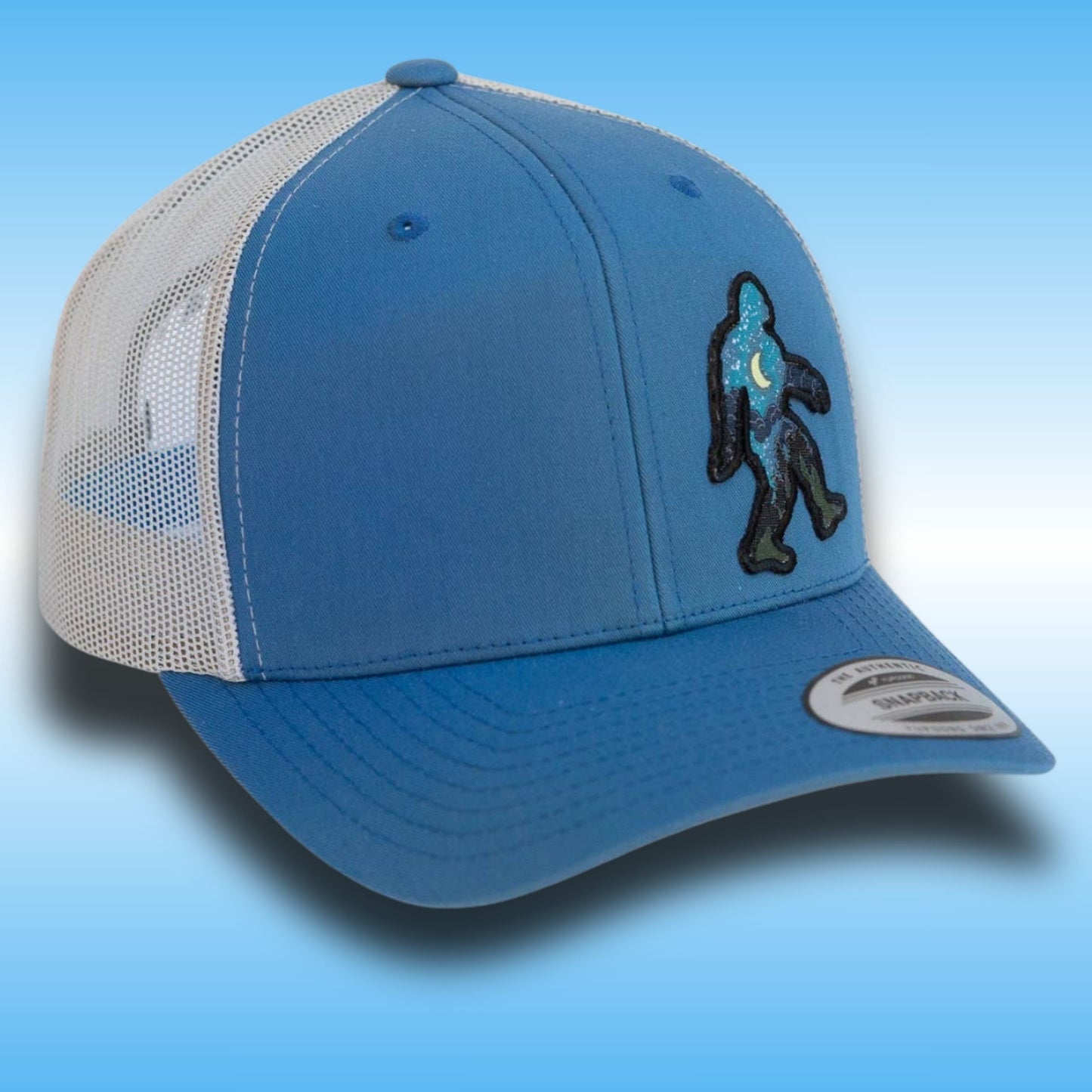 A blue and white trucker hat with a blue front panel, silver mesh back, and a black and blue Bigfoot patch on the side, set against a blue gradient background.