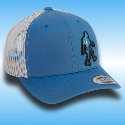 A blue and white trucker hat with a blue front panel, silver mesh back, and a black and blue Bigfoot patch on the side, set against a blue gradient background.