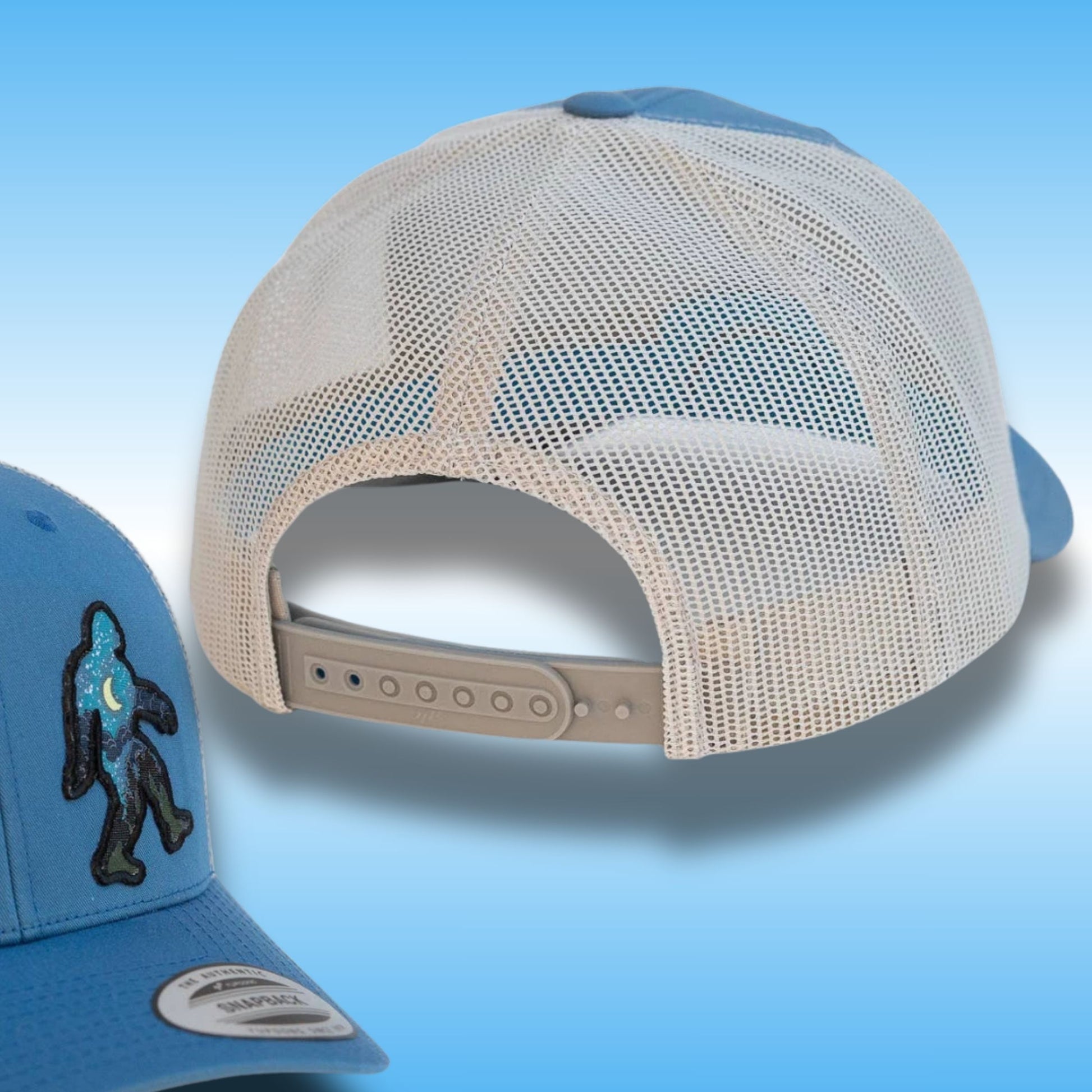 A light blue and silver trucker cap shown from the back, with a smaller inset view of the front-left corner displaying a black embroidered Bigfoot patch on a blue fabric. The cap features a cream mesh back and a light blue structured front and visor.