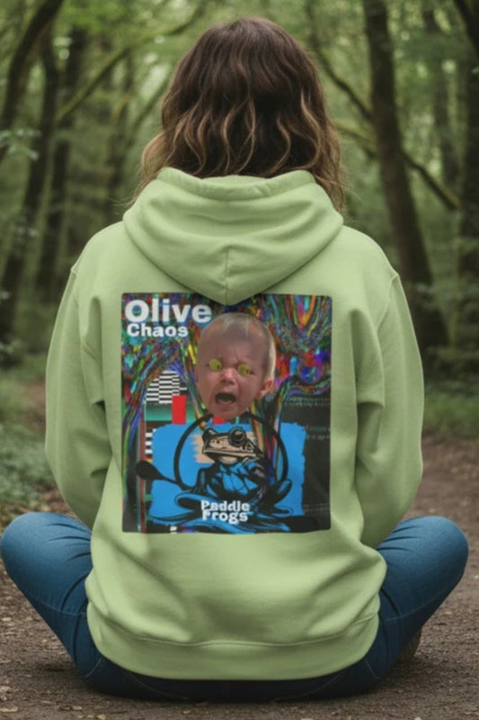 A person sitting cross-legged on a forest path, viewed from behind, wearing a light olive green hoodie with a large graphic print on the back. The graphic features a child's face and the text 'Olive Chaos' and 'Paddy's Frog'.