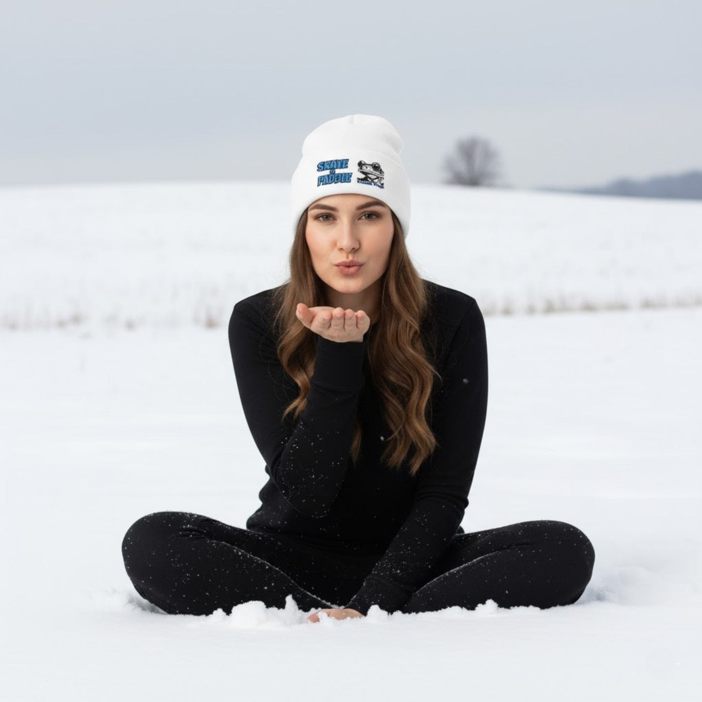 Woman blowing a kiss in the snow wearing a white beanie with a logo. Original Skate & Paddle Beanie Cap by Paddle Frogs white sitting crisscross applesauce in the snow