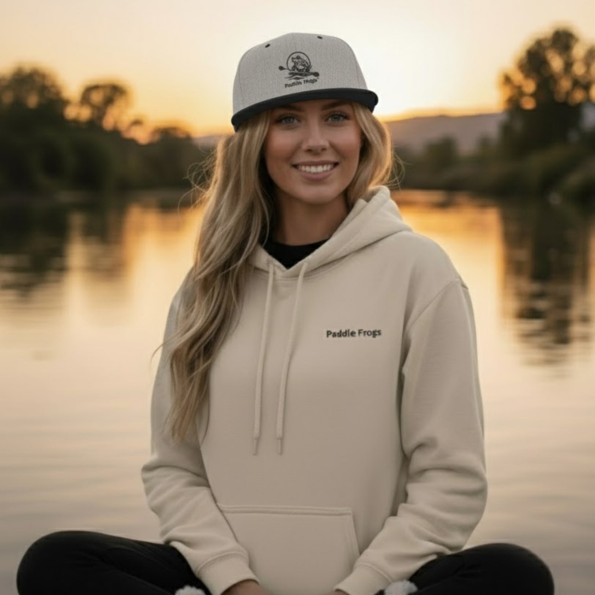 Woman wearing a beige hoodie and cap with 'Paddle Frogs' logo by a lake at sunset.