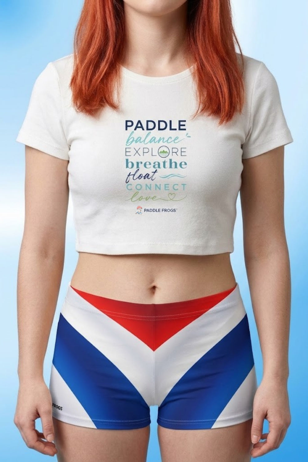 Paddle & Balance Women’s Crop Top | Paddle Frogs™ color White SUP paddleboarding kayaking ladies shirt modeled by Sarah. Also, wearing matching red, white, and blue athletic shorts against a light blue gradient background.