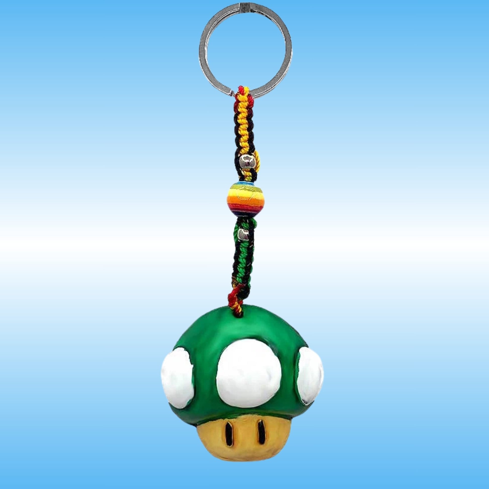 A Super Mushroom keychain featuring a green plastic mushroom with white spots and a gold mouth, attached to a braided cord with red, yellow, and green threads, a striped bead, and a silver metal key ring, set against a blue gradient background.