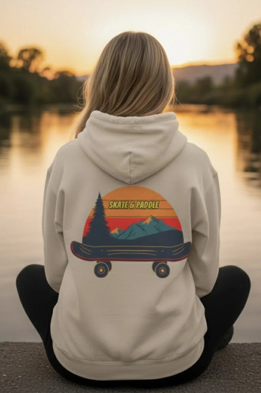 A person sitting cross-legged by a lake at sunset, viewed from behind, wearing a light beige hoodie Chill Mountain Fleece Hoodie  Skate & Paddle Frogs™ with a large graphic on the back showing a skateboard, mountains, trees, and the words 'SKATE ON PADDLE'.